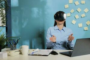 femelle entreprise employé avec virtuel réalité des lunettes à visualiser travail projet. professionnel femme avec immersif multimédia téléspectateur travail à Bureau bureau faire des gestes avec mains. photo