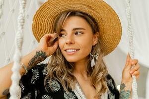 sourire Jeune femme dans boho robe posant dans élégant chambre à coucher. paille chapeau, boho plume laissez tomber des boucles d'oreilles . Dame séance sur pendaison balançoire. photo