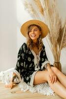 en riant blond femme dans paille chapeau et boho robe posant dans studio plus de blanc Contexte avec pampa herbe décor. oreillers avec macramé ornement. été les tendances. photo