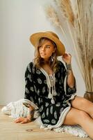 jolie blond femme dans paille chapeau et boho robe posant dans studio plus de blanc Contexte avec pampa herbe décor. oreillers avec macramé ornement. été les tendances. photo