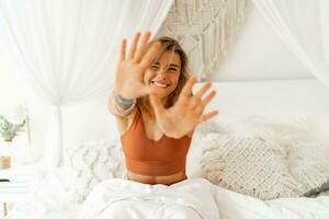 souriant Jeune femme élongation dans lit veille en haut dans confortable chambre avec bohémien intérieur style. femelle portant dans confort pyjamas profiter de bonne heure matin,. photo