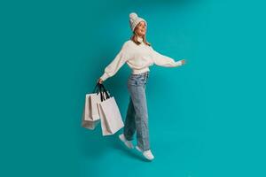 studio hiver portrait de souriant blond femme dans blanc chapeau et laine chandail une en portant achats Sacs , posant dans studio plus de turquose Contexte. plein longueur. photo