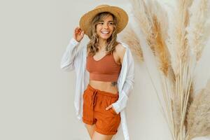 boho ambiance. élégant femme dans été tenue avec paille chapeau posant plus de blanc Contexte dans studio avec pampa herbe décor. photo