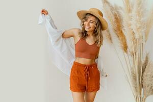 boho ambiance. élégant femme dans été tenue , paille chapeau et Orange vêtements posant plus de blanc Contexte dans studio avec pampa herbe décor. photo