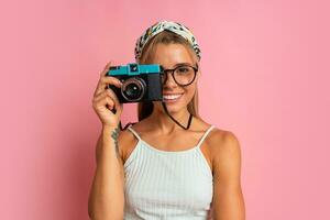en riant blond femme en portant rétro caméra et posant sur rose Contexte. Voyage et été vacances concept. photo
