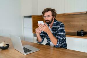 européen homme travaux de maison. il est assis dans le cuisine à le table et les usages une portable ordinateur et parle sur le téléphone. photo