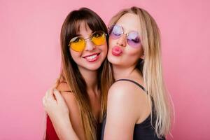 proche en haut studio portrait de deux espiègle Dames étreindre et ayant amusement ensemble sur rose Contexte. brunette femme avec Candide sourire avec sa ami . photo