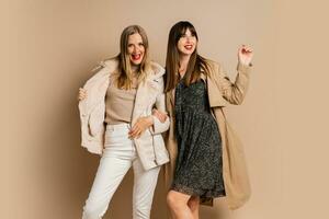 deux élégant femmes dans élégant hiver tenue posant plus de beige Contexte. portant manteau et veste. achats et mode concept. photo