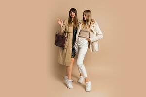 deux élégant femmes dans élégant hiver tenue posant plus de beige Contexte. portant manteau et veste. achats et mode concept. plein longueur. photo