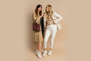 deux élégant femmes dans élégant hiver tenue posant plus de beige Contexte. portant manteau et veste. achats et mode concept. plein longueur. photo