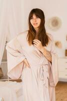 sensuel brunette femme posant dans beige soie kimono dans élégant lumière chambre dans bohème style. photo