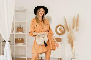 jolie blond femme dans beige lin robe posant dans élégant boho intérieur. photo