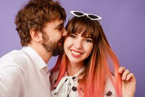 jolie femme avec Candide sourire et rose Cheveux posant avec sa copain avec barbe. branché couple dans l'amour sur violet Contexte. ancien des lunettes de soleil. photo