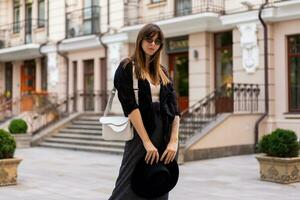 sur de soi élégant brunette femme posant dans européen ville, en marchant sur le rue. portant décontractée tenue, noir chapeau et chemisier. l'automne mode regarder. photo