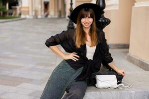 sur de soi élégant brunette femme posant dans européen ville, en marchant sur le rue. portant décontractée tenue, noir chapeau et chemisier. l'automne mode regarder. photo