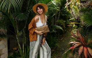 tropical vacances et vacances concept. élégant élégant fille dans blanc Haut et paille chapeau posant sur paume feuilles Contexte dans Bali. photo