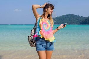 content de bonne humeur Jeune femme écoute à charmant la musique dans écouteurs, en marchant sur le ensoleillé paradis tropical plage. turquoise l'eau Contexte. photo