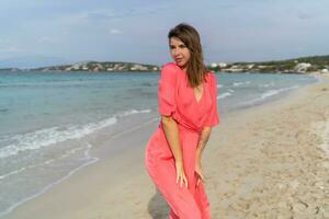 séduisant brunette femme avec tatoo sur main dans rose robe posant sur le plage. photo