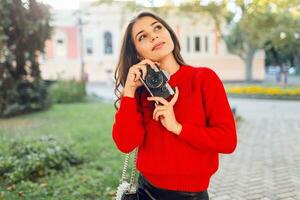 ensoleillé mode de vie image de jolie brunette fille dans rouge décontractée arrêtez-vous et jupe fabrication des photos par photo caméra dans ensoleillé parc. en marchant dans ville jardin et à la recherche sites touristiques .