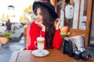 étourdissant Jeune Dame dans élégant noir chapeau et brillant rouge chandail séance dans ouvert espace café et boisson café avec Lait ou cappuccino. photo