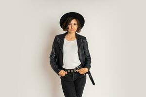 studio mode photo de élégant brunette femme dans noir chapeau et cuir veste posant sur blanc antécédent. blanc T-shirt.