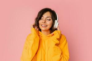 bienheureux souriant femme dans blanc écouteurs écoute à la musique sur rose Contexte. portant Orange sweatshirt à capuche. photo