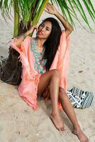 été portrait de Jeune magnifique asiatique femme relaxant sur tropical plage, portant boho branché robe, séance sur blanc le sable près paume arbre. mode regarder. photo