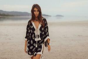 magnifique asiatique femme dans tropical bohémien robe posant sur le plage. portant des boucles d'oreilles avec plume, Collier et bracelets. photo