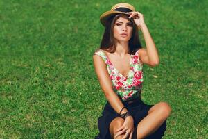 proche en haut Extérieur portrait de séduisant Jeune femme souriant , séance sur vert Frais herbe dans ensoleillé printemps parc . parfait bronzer peau. longue svelte jambes. photo