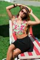 proche en haut Extérieur portrait de séduisant Jeune femme souriant , séance sur vert Frais herbe dans ensoleillé printemps parc . parfait bronzer peau. longue svelte jambes. photo