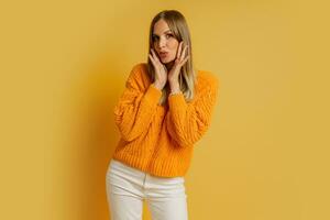 jolie blond femme avec surprise visage dans Orange élégant l'automne chandail posant plus de Jaune Contexte dans studio. photo