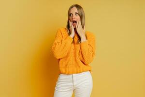 jolie blond femme avec surprise visage dans Orange élégant l'automne chandail posant plus de Jaune Contexte dans studio. photo