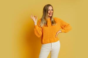 content blond femme dans Orange élégant l'automne chandail posant plus de Jaune Contexte dans studio. montrant signe d'accord. photo