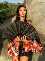 asiatique femme dans coloré soie kimono chemisier en portant noir ventilateur et posant sur le plage avec rochers. photo