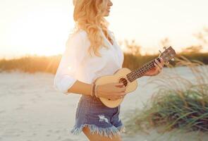 galbé gingembre Dame en jouant ukulélé guitare dans Soleil lumière sur le plage . portant paille chapeau et branché jeans shorts. chaud le coucher du soleil couleurs. photo