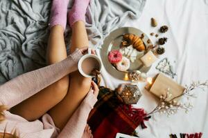 Haut vue image de Jeune svelte femme profiter Matin temps dans sa lit, portant chaud confortable de laine chandail et rose chaussettes, en portant gros tasse de café. photo