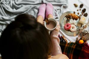 Haut vue image de Jeune svelte femme profiter Matin temps dans sa lit, portant chaud confortable de laine chandail et rose chaussettes, en portant gros tasse de café. photo