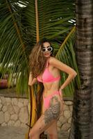 magnifique élégant femme dans branché rose maillots de bain posant sur le plage. été vacances, tropical île, palmiers. portant décontractée des lunettes de soleil. photo