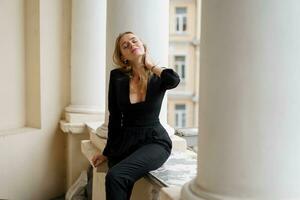 blond femme dans noir tenue posant sur terrasse. portrait de content fille je souriant et repos sur balcon. photo