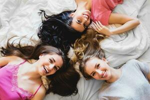 Haut vue de Trois les filles dans lit . groupe de jolie Jeune femmes dans pyjamas mensonge sur retour et parler. ensoleillé chaud couleurs. confortable atmosphère photo