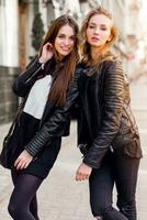 deux élégant jolie les filles posant sur le rue dans ensoleillé journée. portant branché Urbain tenue , cuir veste et bottes talons. photo