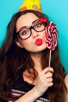 fou marrant jolie femme posant contre bleu mur portant branché été tenue , cool mignonne des lunettes et fête chapeau. en portant gros rose sucette . photo