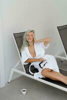 magnifique blond femme dans blanc peignoir effrayant sur plate-forme chaise dans luxe Hôtel. enjoing Matin et en buvant café. photo