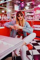 à la mode fille dans jeans veste séance dans t rétro américain café. charmant souriant femme profiter sucré Lait secouer . portant blanc baskets. photo