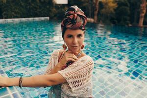 femme avec brillant faire en haut dans bohémien tenue, turban, ethnique des boucles d'oreilles, Collier et bracelets posant dans nager bassin. photo
