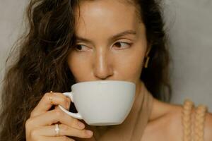 proche en haut portrait de magnifique brunette femme avec parfait ondulé Cheveux profiter tasse de café dans moderne café. photo