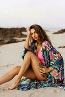 portrait de magnifique brunette femme dans sexy maillots de bain et boho citron avec tropical impression posant sur le plage dans le coucher du soleil lumière. parfait ondulé Cheveux, bronzer parcourir corps. photo