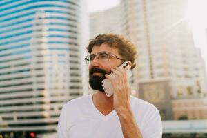 Beau homme avec barbe parlant par mobile téléphone tandis que en marchant dans gros moderne ville avec grattes ciels. en plein air à le coucher du soleil . portant blanc t chemise. photo