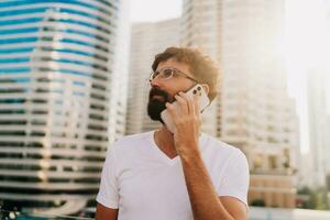 Beau homme avec barbe parlant par mobile téléphone tandis que en marchant dans gros moderne ville avec grattes ciels. en plein air à le coucher du soleil . portant blanc t chemise. photo