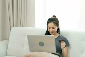femme asiatique tenant une carte de crédit pour faire des achats en ligne tout en étant assise sur le canapé pendant les vacances à la maison. photo